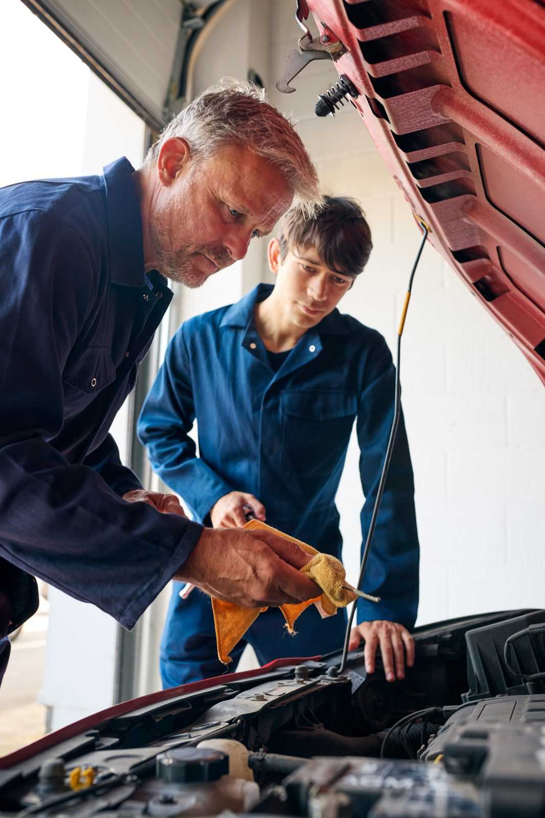 Male Car Auto Mechanic With Trainee Looking Under Bonnet Of Car Checking Oil Level With Dipstick